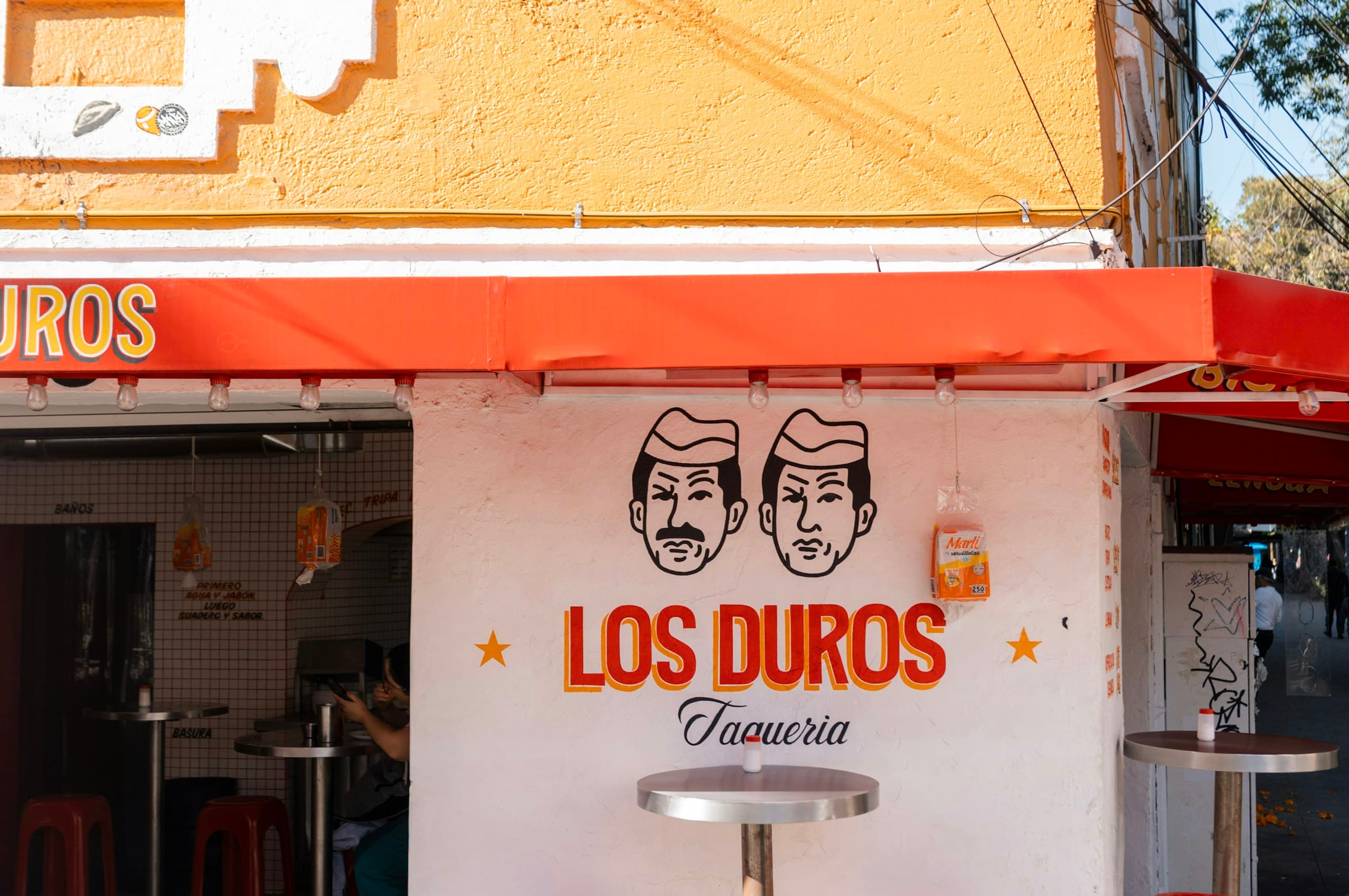Taquería de esquina con mesas de acero y bancos rojos en la banqueta